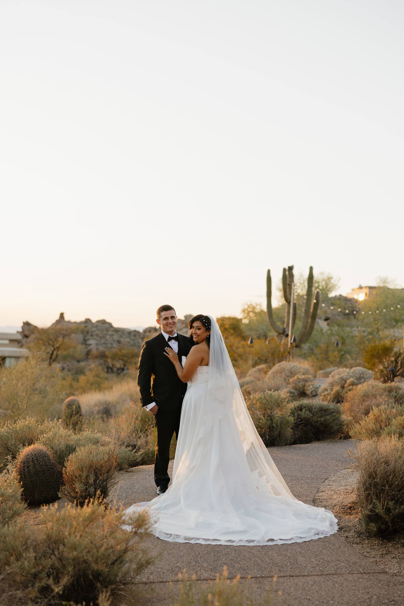 A bride and groom on a desert path at dusk, her train spread behind her