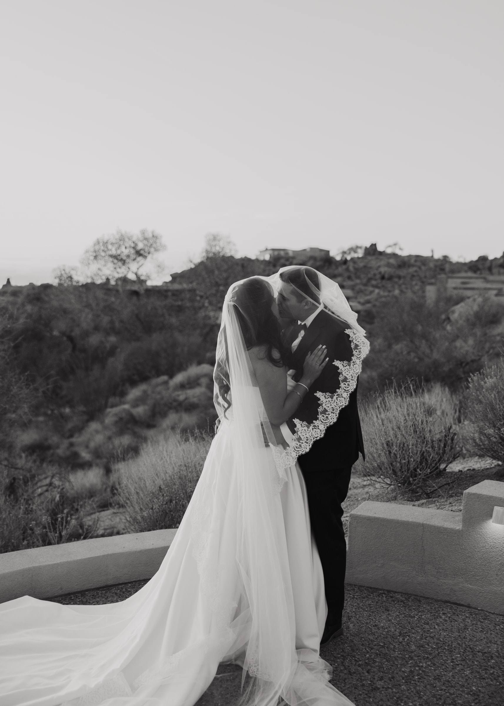 A bride and groom kissing under her veil, its long train spread on the terrace, in black and white