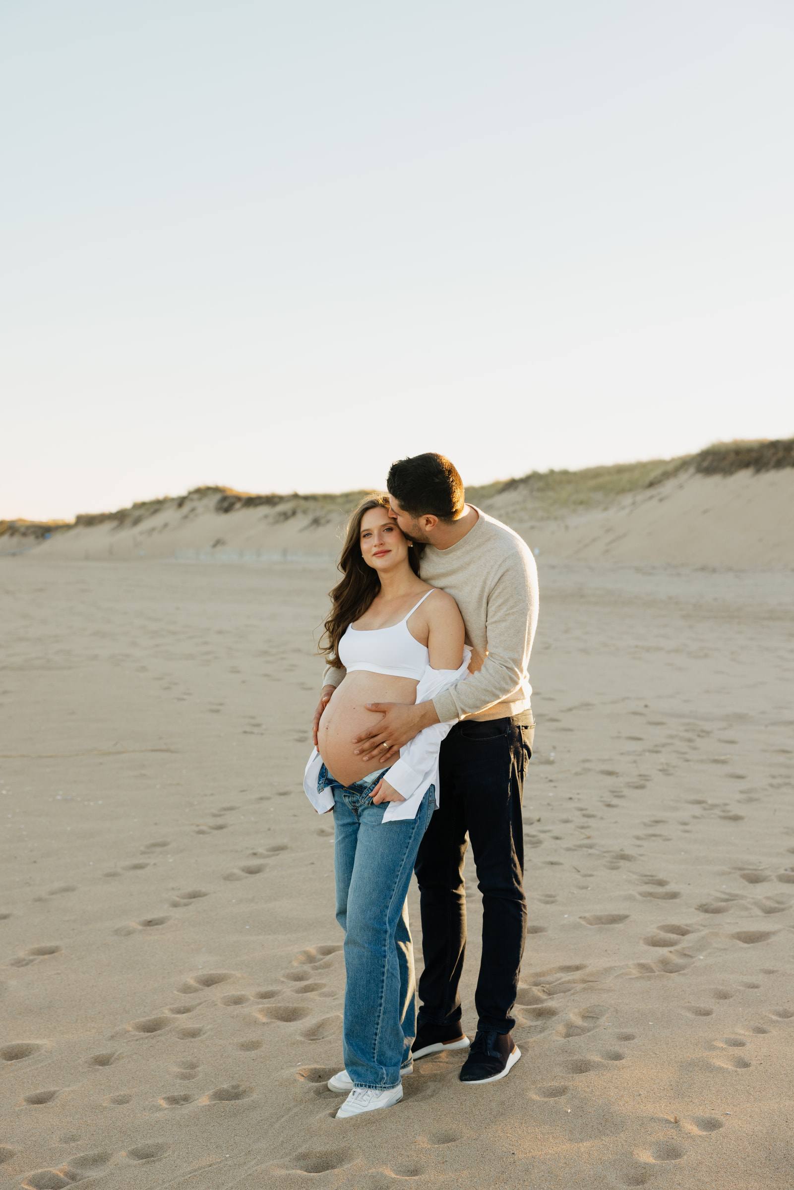 An expecting couple embracing on a sandy beach at golden hour