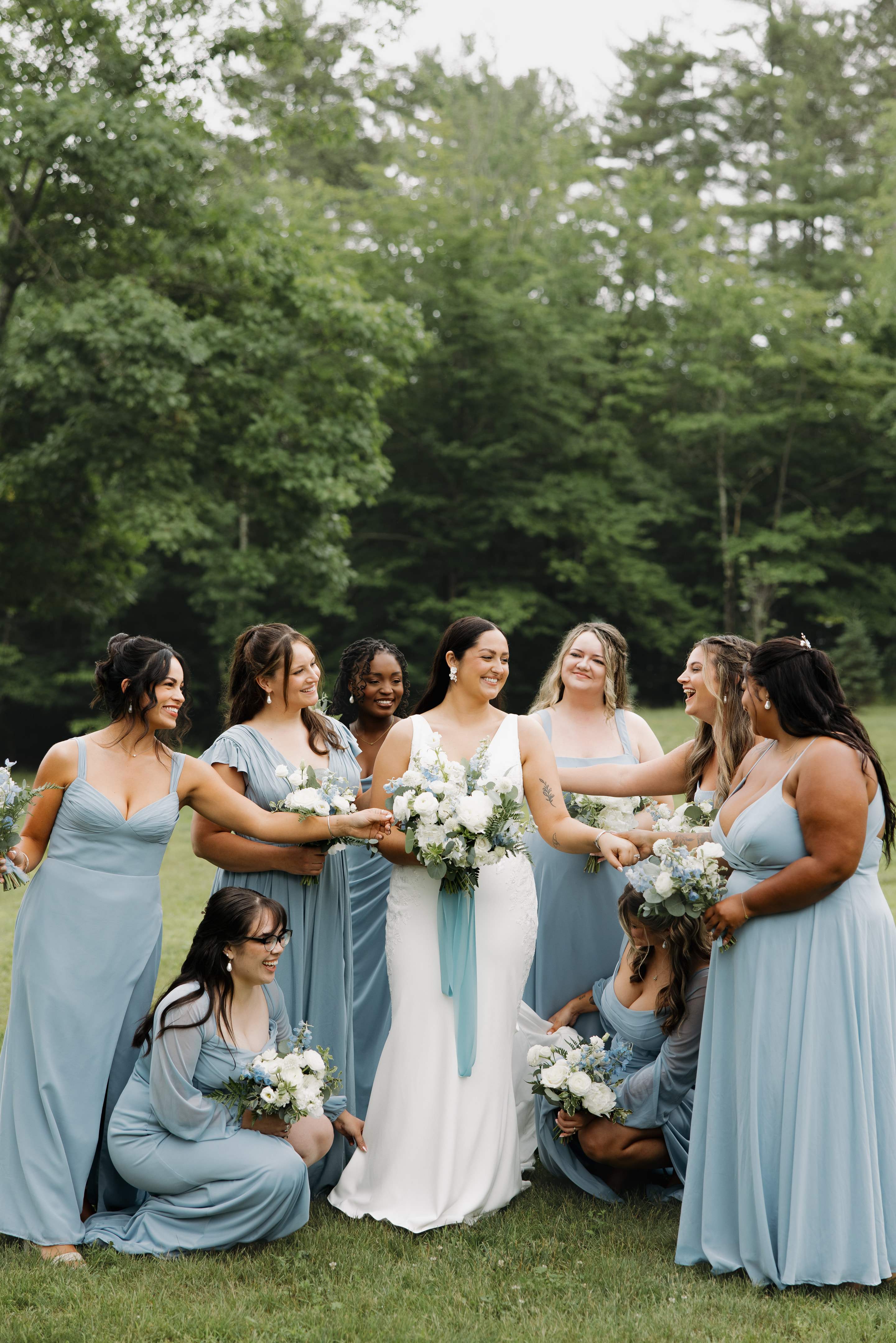 A laughing bride holding hands with her bridesmaids in dusty-blue dresses on a lawn