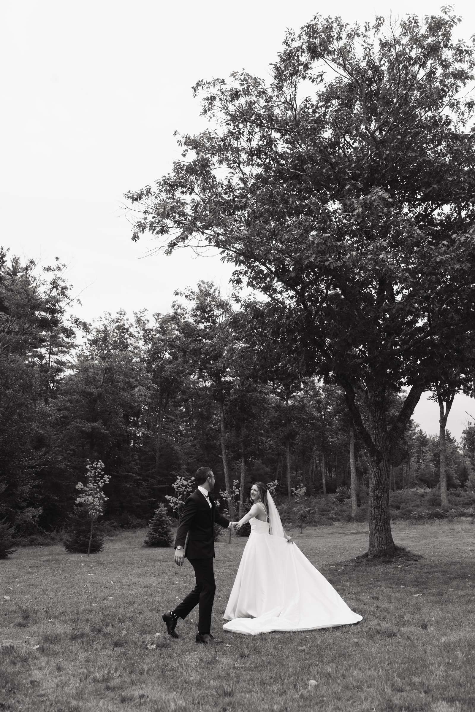 A bride and groom walking hand in hand across a field beneath a tall tree, in black and white