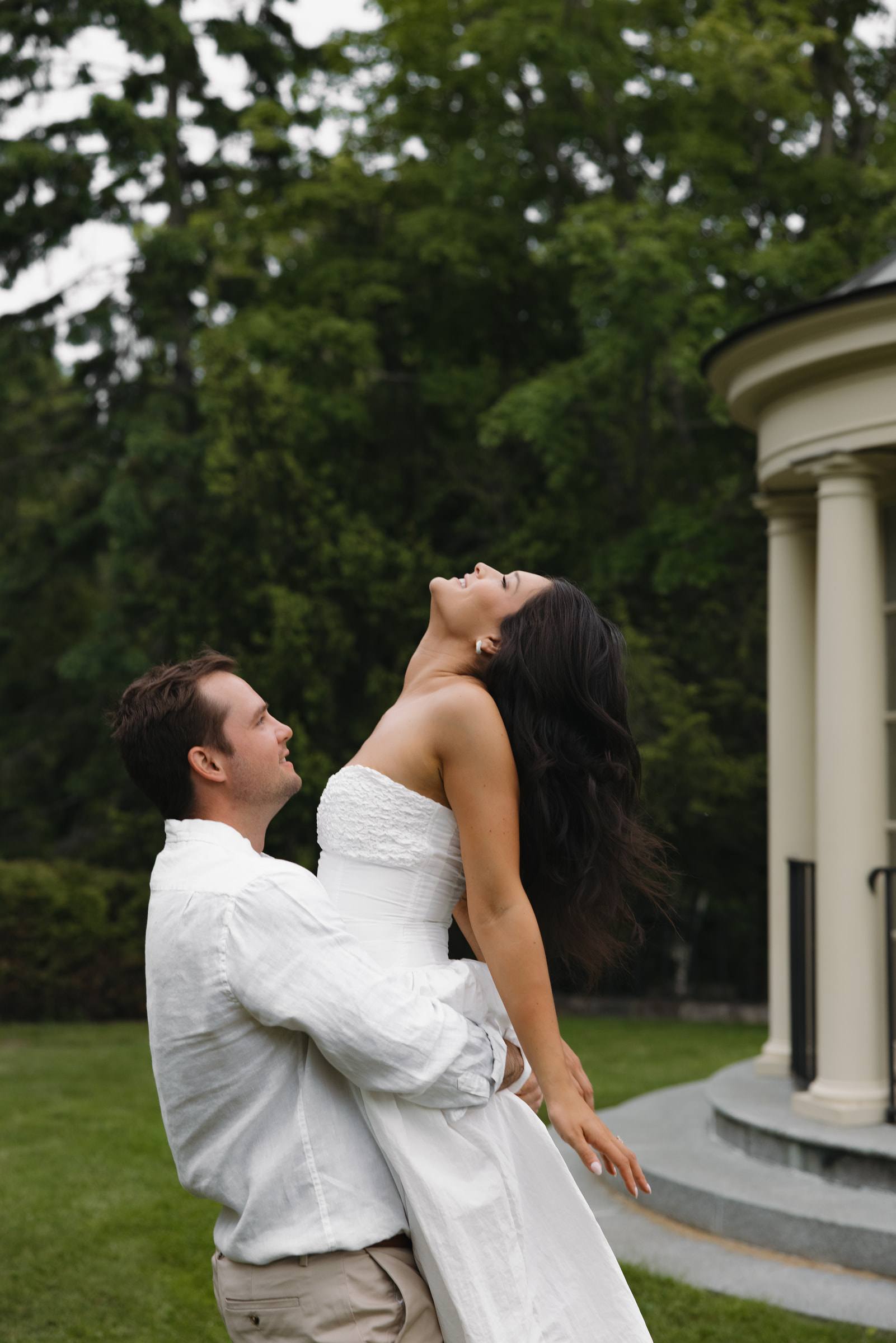 A groom lifting his laughing bride beside a white garden rotunda