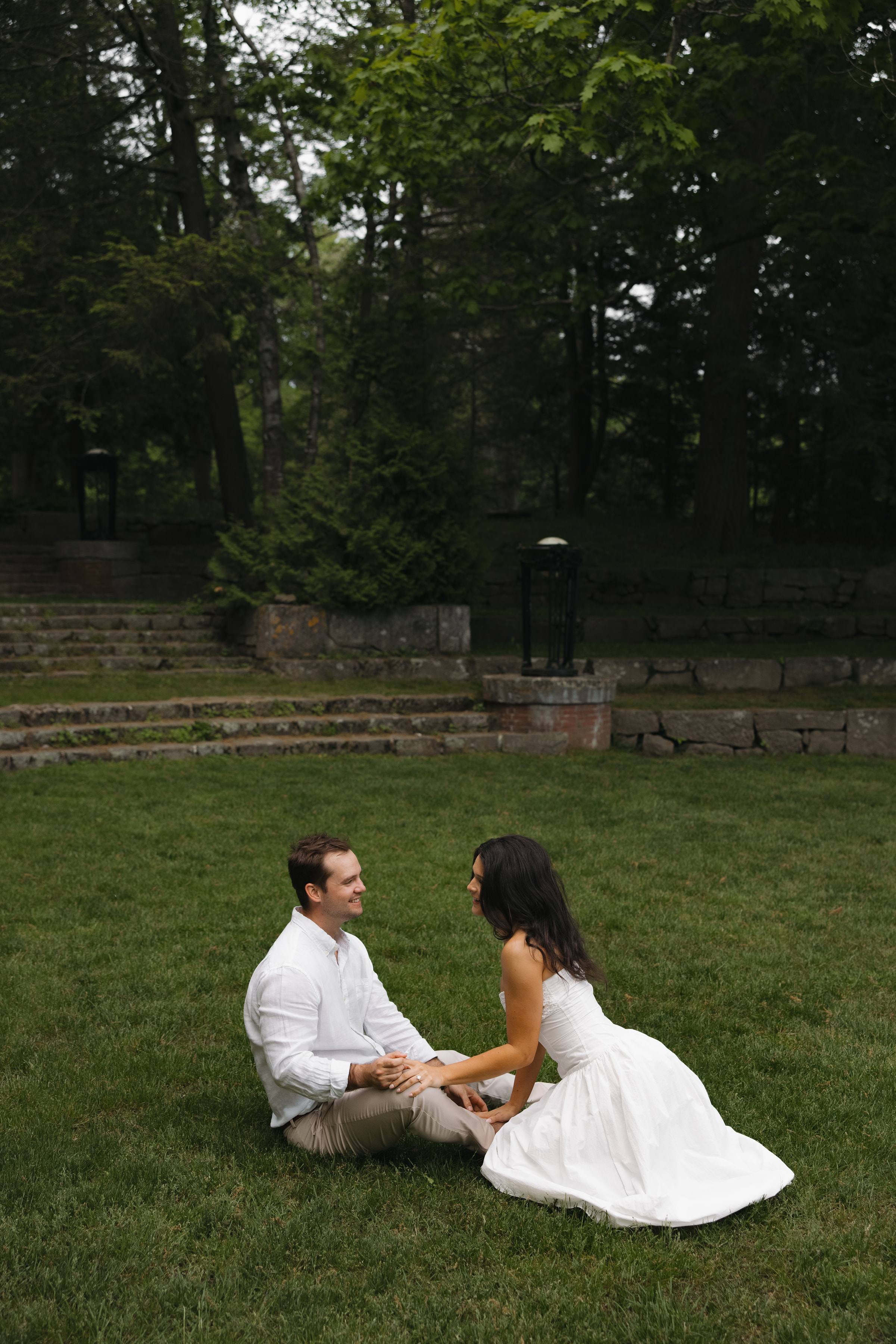 A couple sitting together on a lawn below a line of dark trees