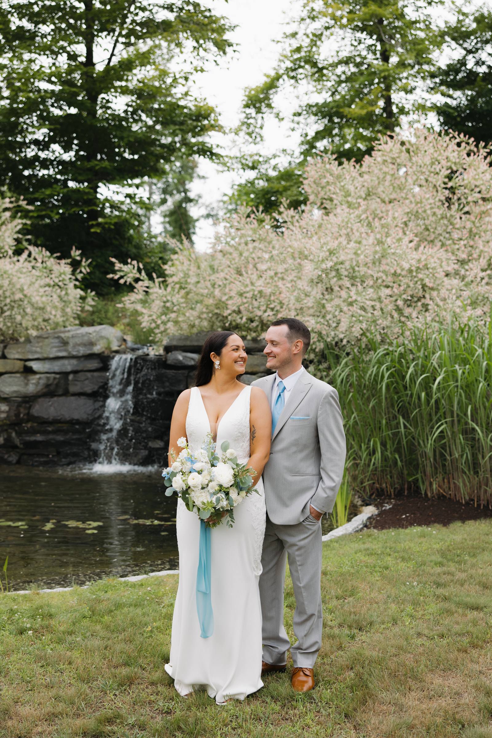 A bride and groom smiling at each other beside a garden waterfall