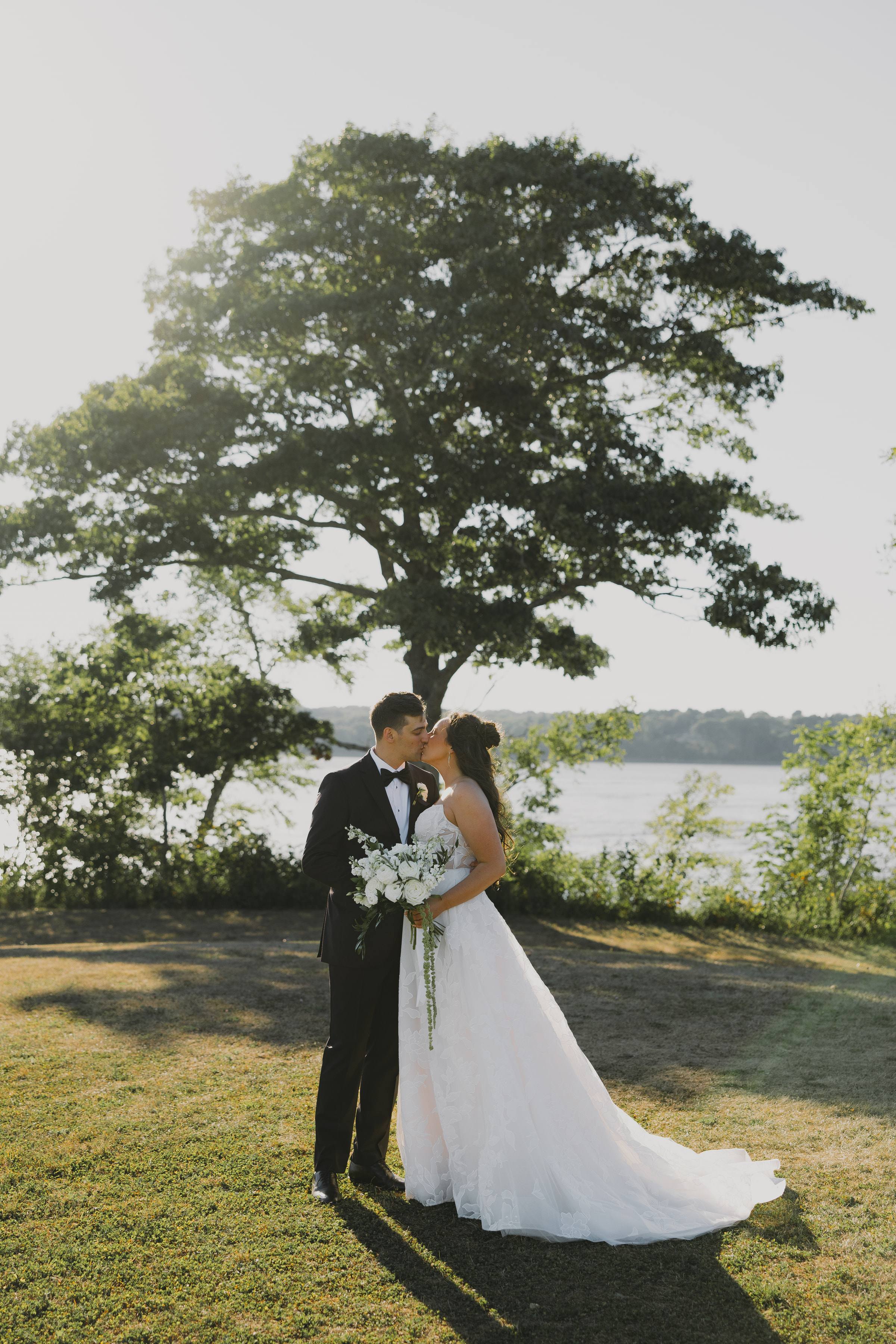 A bride and groom kissing beneath a tree beside a lake