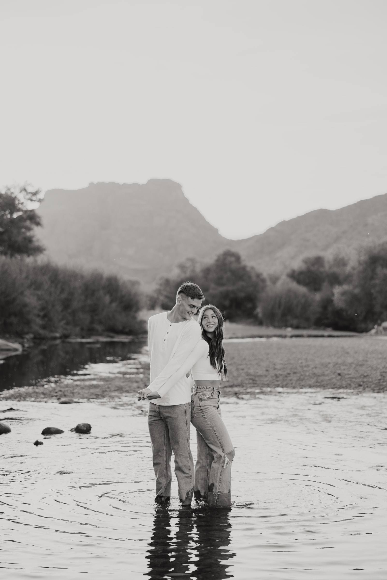 A couple standing together in a shallow desert river below a mesa, in black and white