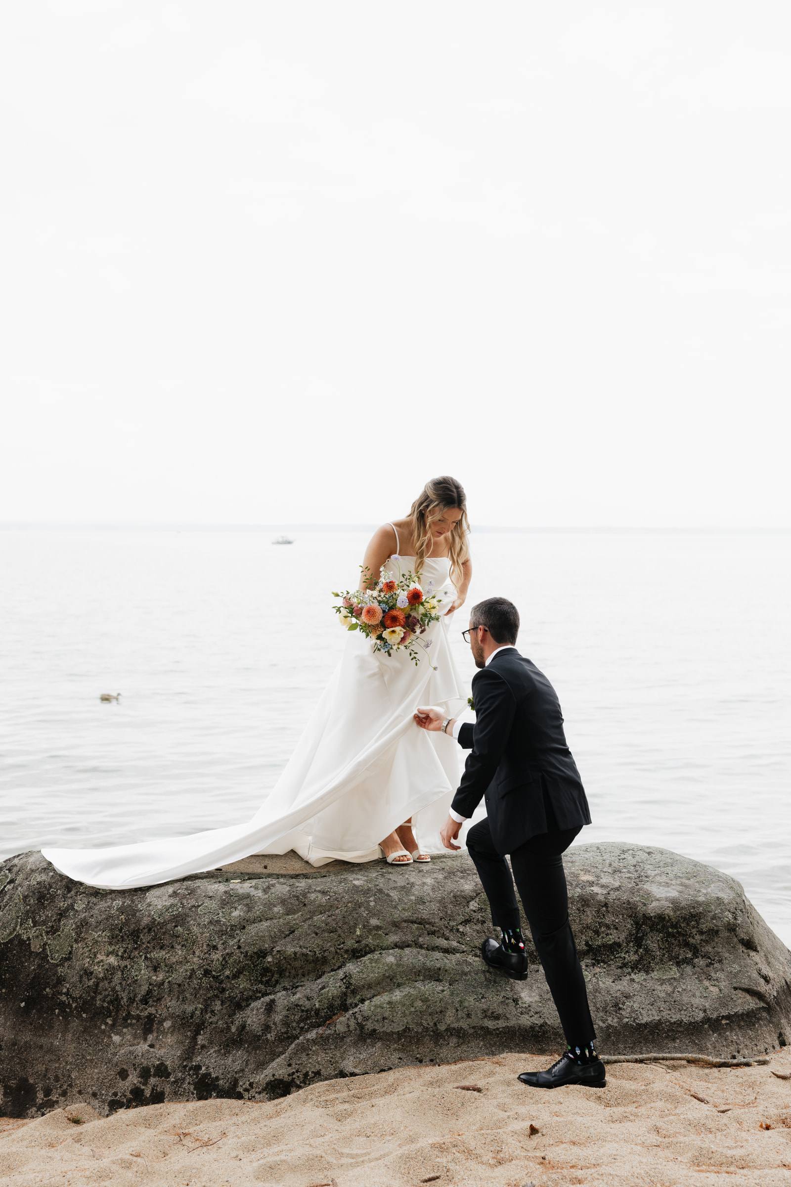 A groom helping his bride climb onto a rock at the edge of the sea
